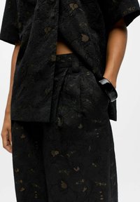 Schwarzes, blumendekoriertes Zweiteiler-Outfit mit einem locker geschnittenen Oberteil und weit geschnittenen Hosen. Strukturiertes Gewebe mit dezenten goldenen Akzenten.