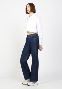 Gekürztes weißes Hemd mit Kragen, kombiniert mit dunkelblauen weiter geschnittenen Jeans. Die Sneakers sind grau mit weißen Akzenten.