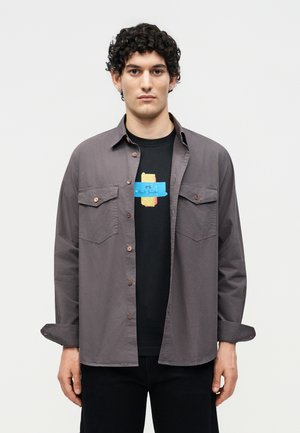 Νέος άνδρας με σγουρά μαλλιά που φορά γκρι πουκάμισο με κουμπιά πάνω από μαύρη γραφική μπλούζα, στέκεται μπροστά σε απλό λευκό φόντο.