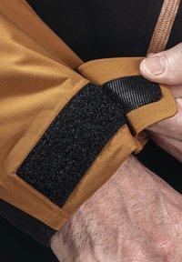 Brauner Stoff mit glatter Oberfläche, ausgestattet mit einem schwarzen Klettverschluss-Patch und strukturierten Akzenten am Bündchen. Hand wird gezeigt, die den Ärmel hält.