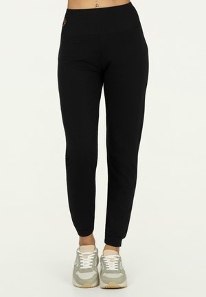 Vrouw die zwarte hoge taille slim-fit joggerbroek en witte grijze sneakers draagt, staand tegen een effen lichte achtergrond.