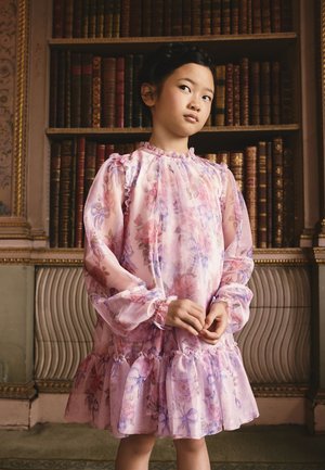 Jeune fille en robe fleurie rose se tient les mains jointes devant une grande étagère en bois remplie de vieux livres.