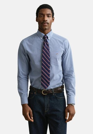 Mężczyzna w niebieskiej koszuli w kratę na guziki, granatowym krawacie w paski, ciemnych dżinsach i brązowym plecionym pasku, stojący na jednolitym tle.