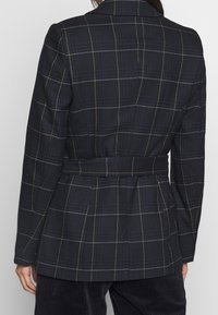Granatowa, kratkowana marynarka z paskiem, wykonana z teksturowanej tkaniny z kontrastującymi białymi i złotymi liniami w kratkę. Kołnierz z wycięciem i dopasowany krój.