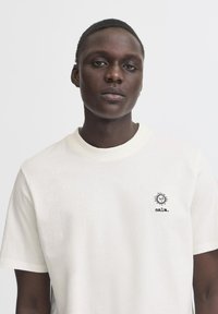 Krämfärgad bomull t-shirt med rund hals, korta ärmar, med en liten svart sol-logotyp och ordet "calm." broderat på vänster bröst.
