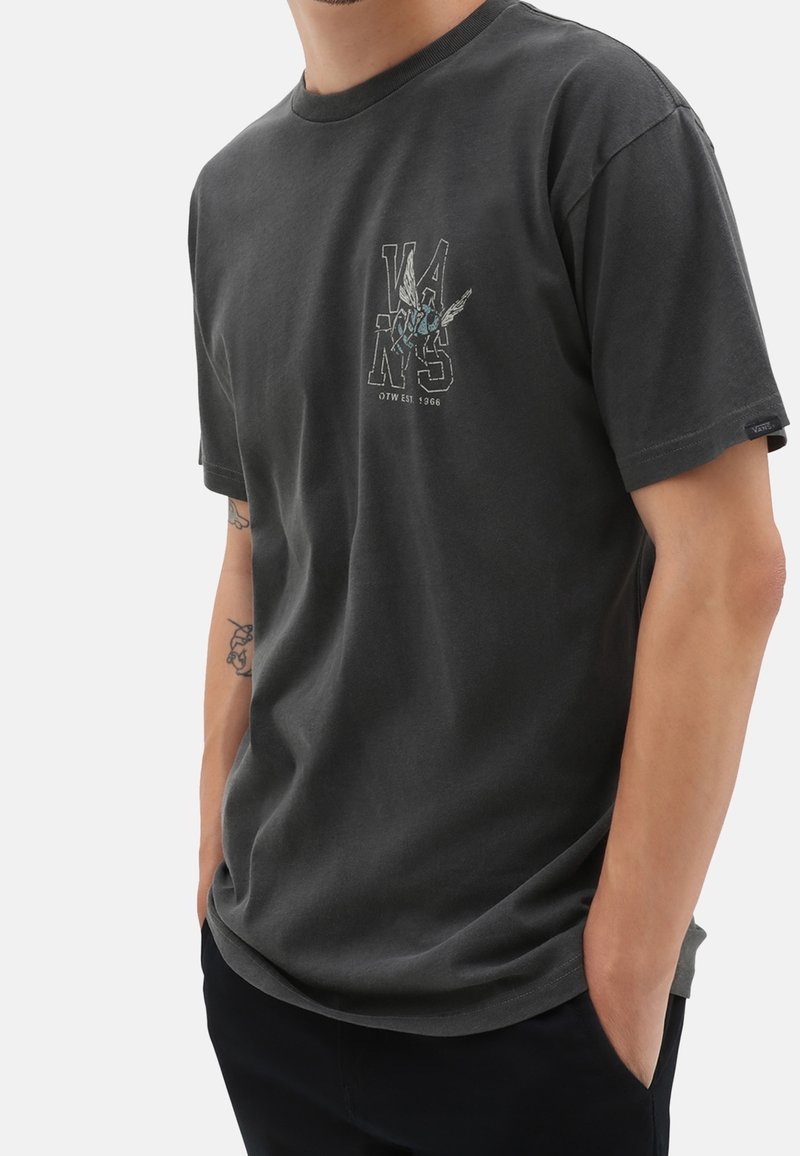 Homme portant un t-shirt Vans gris foncé avec un petit graphisme et texte sur la poitrine gauche, debout les mains dans les poches.