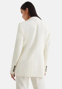 Blazer bianco su misura con colletto a scollo, una fessura sul retro e dettagli in bottone nero sui polsini. Texture liscia, design moderno.