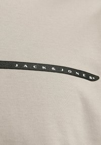 Tejido de camiseta beige claro que presenta una franja gráfica negra brillante con el texto "JACK & JONES" en blanco, centrado horizontalmente.