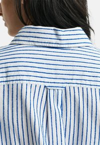 Witte blouse met blauwe strepen, voorzien van een kraag en een plooi aan de achterkant. De stof lijkt textuur te hebben met een casual, ontspannen pasvorm.