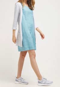 Cardigan leggero, a maniche lunghe e aperto davanti, di colore bianco, indossato sopra un vestito senza maniche di lunghezza al ginocchio in aqua. Scarpe da ginnastica grigie.