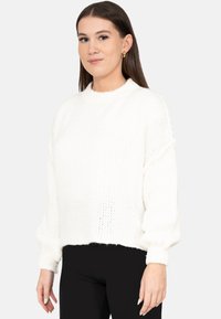 Jonge vrouw met steil bruin haar draagt gouden oorringen, een witte gebreide trui met lange mouwen en een zwarte broek tegen een effen achtergrond.