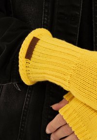 Geel gebreide handschoenen met ribbelstructuur, voorzien van een bruine leren label. Draag over een zwart stoffen kledingstuk.