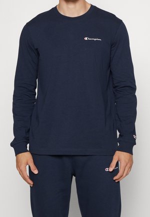 Mann trägt dunkelblaues langärmeliges Champion-Shirt und passende Hose, beide mit kleinen Champion-Logos auf Brust, Handgelenk und Oberschenkel.