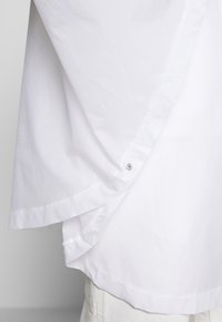 Vit skjorta med en rundad fåll, kontrasterande textur och säkrad med en tryckknapp vid sidsömmen. Slät tygfinish.