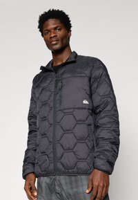 Man wearing a black hexagon-quilted zip-up jacket with a chest pocket and side pockets, standing against a plain light background.
