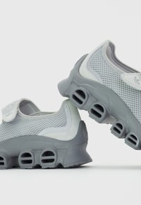 Scarpe sportive grigie realizzate in mesh e suede, dotate di chiusura a strappo, superficie testurizzata e una suola distintiva con ritagli circolari.