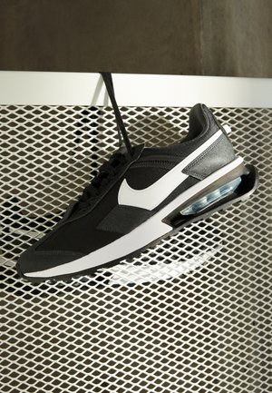 Czarno-biały but sportowy Nike wiszący na sznurowadłach na białej metalowej siatce, podkreślający podeszwę z poduszką powietrzną.