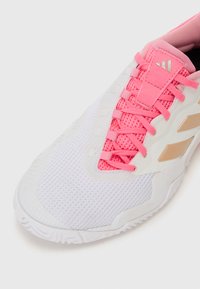 Zapatilla deportiva blanca con material de malla, cordones rosas y detalles en dorado. Presenta una suela texturizada y una lengüeta acolchada para mayor comodidad.