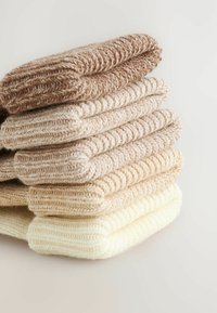 Stapel von fünf gefalteten gestrickten Pullovern in Farbverlauf von Creme bis Hellbraun auf einer weißen Oberfläche.