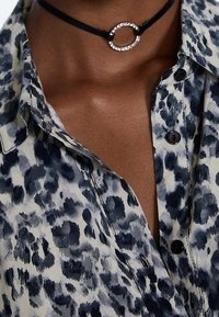 Schwarzes Halsband mit rundem Kristallakzent, kombiniert mit einem blau-weißen Animal-Print-Hemd mit Knopfleiste.