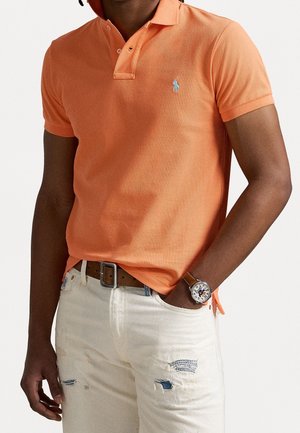 Man draagt een oranje polo met korte mouwen, witte gescheurde spijkerbroek, bruine riem en een polshorloge aan de linkerhand in de zak.