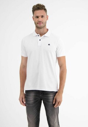 Mann trägt weißes Kurzarm-Poloshirt mit kleinem Logo und dunkel verwaschene Jeans, steht vor einem schlichten weißen Hintergrund.