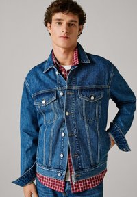 Chaqueta vaquera en azul oscuro, con cierre frontal de botones, cuello clásico, bolsillos en el pecho y una camisa de cuadros roja a contraste debajo.