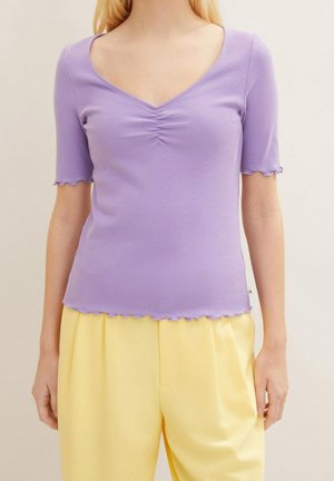 Lila, geripptes Kurzarm-Top mit V-Ausschnitt und Raffdetail an der Brust. Mit geschwungenem Saum und Struktur. Kombiniert mit gelben Hosen.