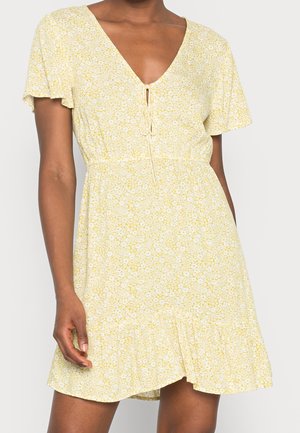 Kobieta w jasnos żółtej sukience w kwiaty z krótkim rękawem, dekoltem w serek i wiązaniem z przodu, stojąca na jednolitym tle.