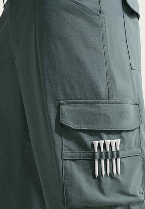 Paliki do golfa schowane w pasku kieszeni zielonych spodni bojowych, podkreślając kontekst przechowywania i aktywności na świeżym powietrzu.