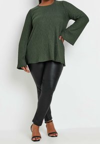 Top verde oliva con texture e maniche a campana lunghe, abbinato a leggings neri in finta pelle e tacchi neri con cinturini.