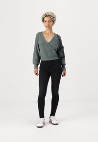 Green knitted wrap sweater with long sleeves, paired with black skinny jeans and white sneakers featuring red and blue accents.