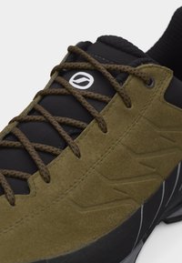 Εγγύς πλάνο ενός παπουτσιού σνίκερ από ελιά πράσινο σουέτ με μαύρη γλώσσα, καφέ κορδόνια με στίγματα και λευκό κυκλικό λογότυπο στη γλώσσα.