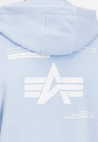 Világoskék kapucnis pulóver, fehér nyomtatott logókkal és szöveggel a hátsó oldalon. Puha anyag, laza design és minták nélkül.