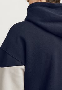 Sudadera con capucha navy con un panel gris claro en el lado inferior y un corte holgado y relajado. Textura suave, con un detalle de costura mínima.