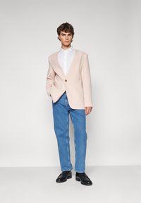 Hellrosa Oversize-Blazer mit einem goldenen Knopf, weißes Hemd, blaue gerade Jeans und schwarze Lederschuhe.