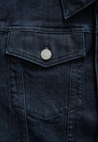 Close-up of dark blue denim jacket pocket with flap secured by a metal button labeled "Premium Denim Goods."