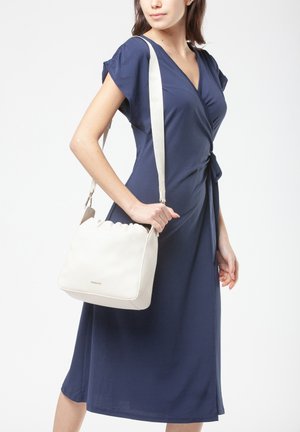 Donna che indossa un vestito avvolgente blu navy che porta una borsa a tracolla bianca con una larga tracolla, in piedi davanti a uno sfondo bianco.