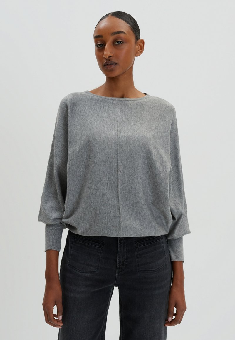 Pullover gris léger avec un col bateau, coupe ample et manches longues. Porté avec un jean noir, affichant un look décontracté et relax.
