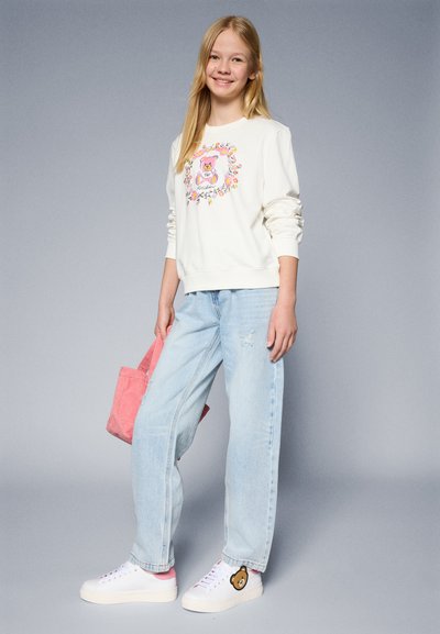 Sudadera blanca con un gráfico de un oso y diseño floral, combinada con jeans azul claro y zapatillas blancas con un parche de oso. Bolsa de tote rosa.