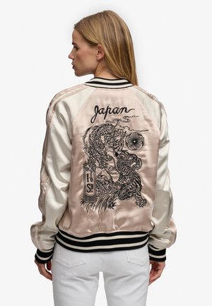 Mujer que lleva una chaqueta bomber de satén con bordado de tigre y dragón en negro y el texto "Japan" en la espalda, combinada con pantalones blancos.
