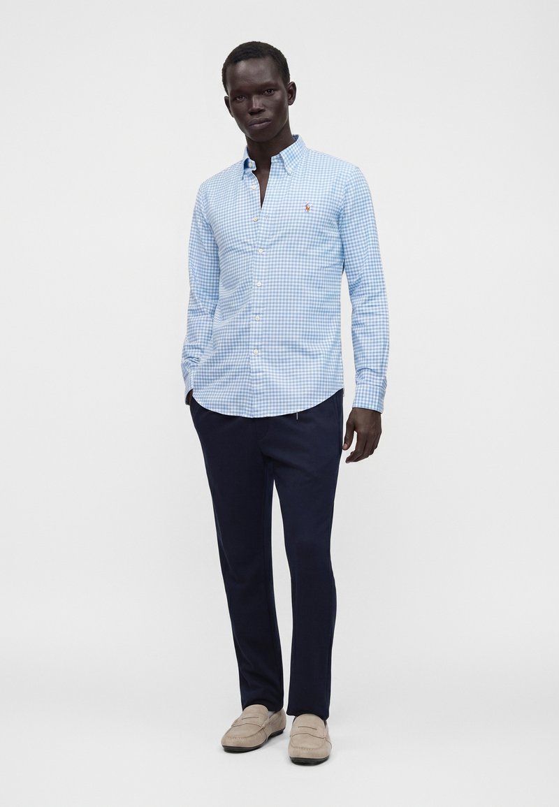 Homme portant une chemise à carreaux bleu clair à boutons, un pantalon bleu marine foncé et des mocassins beige, debout devant un fond blanc uni.