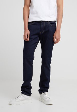 Persoon die donkerblauwe slim-fit jeans draagt met oranje stiksels en witte sneakers, staand tegen een effen lichte achtergrond.