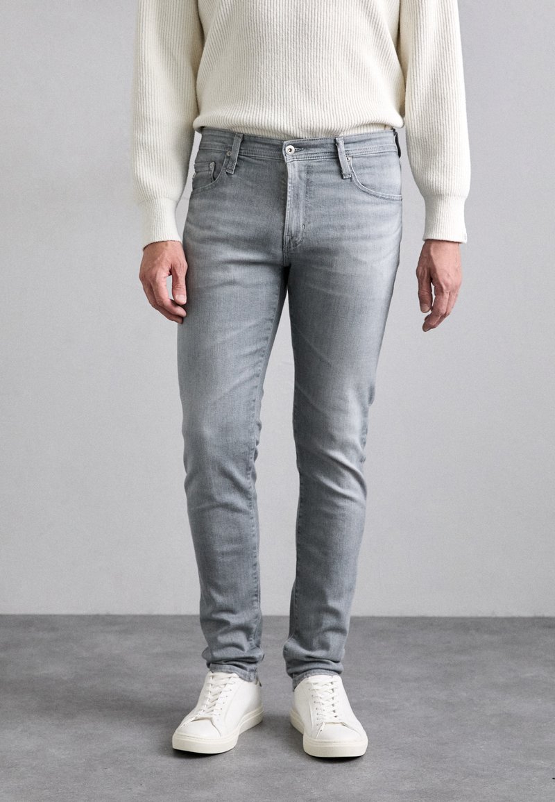 Gray skinny jeans made of denim, featuring a mid-rise waist, five pockets, and subtle fading. Paired with white sneakers.