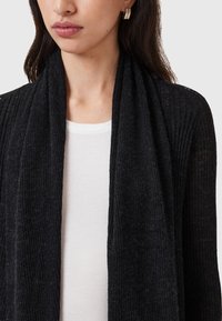 Black ribbed cardigan made of lightweight fabric, featuring an open front design. Worn over a white textured top, with subtle ear accessories.
