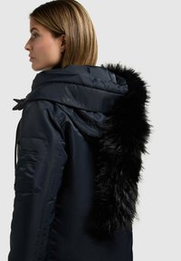 Giubbotto puffer blu navy con collo in pelliccia sintetica nera staccabile, dotato di tasca con zip sulla manica e cinturini regolabili per il cappuccio.