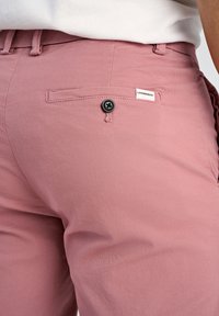 Pantaloni in cotone rosa con una texture liscia, caratterizzati da una tasca posteriore con un bottone nero e un piccolo etichetta bianca cucita sulla vita.