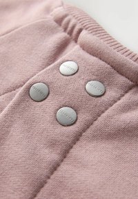 Hele roosa kangas, millel on tekstuuriga pind ja neli metallist nööbi, millel on silt "ALLSAINTS", paigutatud vertikaalselt.