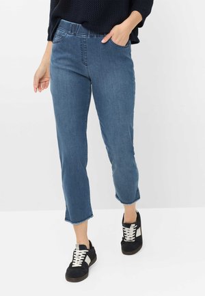 Blå cropped jeans med frynsete kanter, båret med sorte og hvide sneakers, hånd afslappet i lommen, mørk tekstureret sweater synlig over taljen.