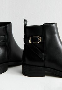 Botas negras de tobillo con textura de cuero liso, cremallera lateral, tira elástica y acento de hebilla en tono dorado en el lateral. Suela plana y estriada.